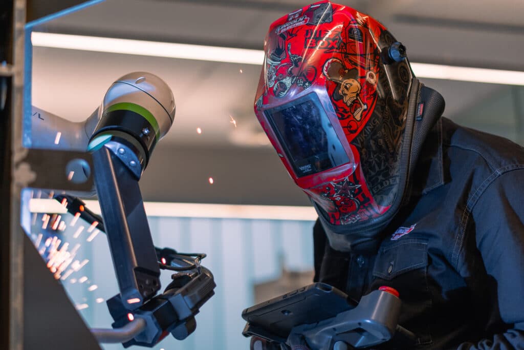 Operator in a welding helmet collaborating with a robotic welding arm, sparks flying as the robot performs precise welding tasks. A clear example of human-robot teamwork in manufacturing.